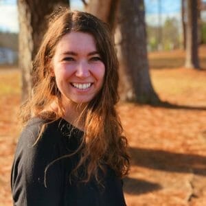 Photo of artist Lindsay Holifield, taken outside in the autumn. Lindsay has pale skin, blue eyes, and long brown hair that is a bit wind-swept in the photo. Her smile is infectious.